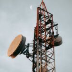 Vertical shot of a telecommunication tower with dishes under a cloudy sky and visible moon.