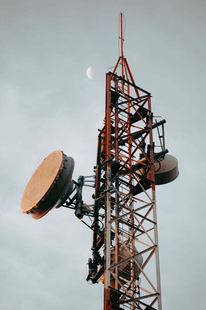 gallery-5 Vertical shot of a telecommunication tower with dishes under a cloudy sky and visible moon.