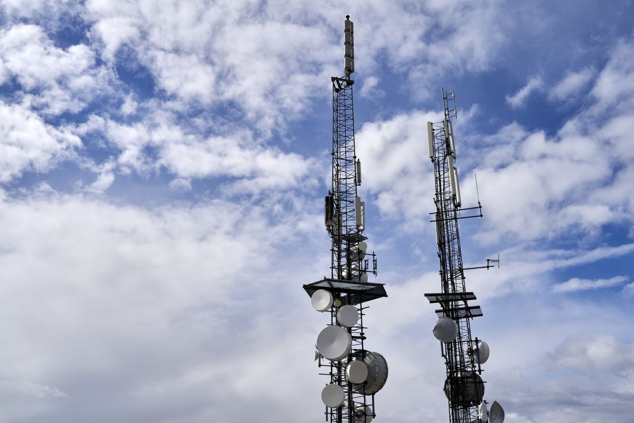 hero-img-02 Telecommunication towers with antennas and dishes under a bright cloudy sky.